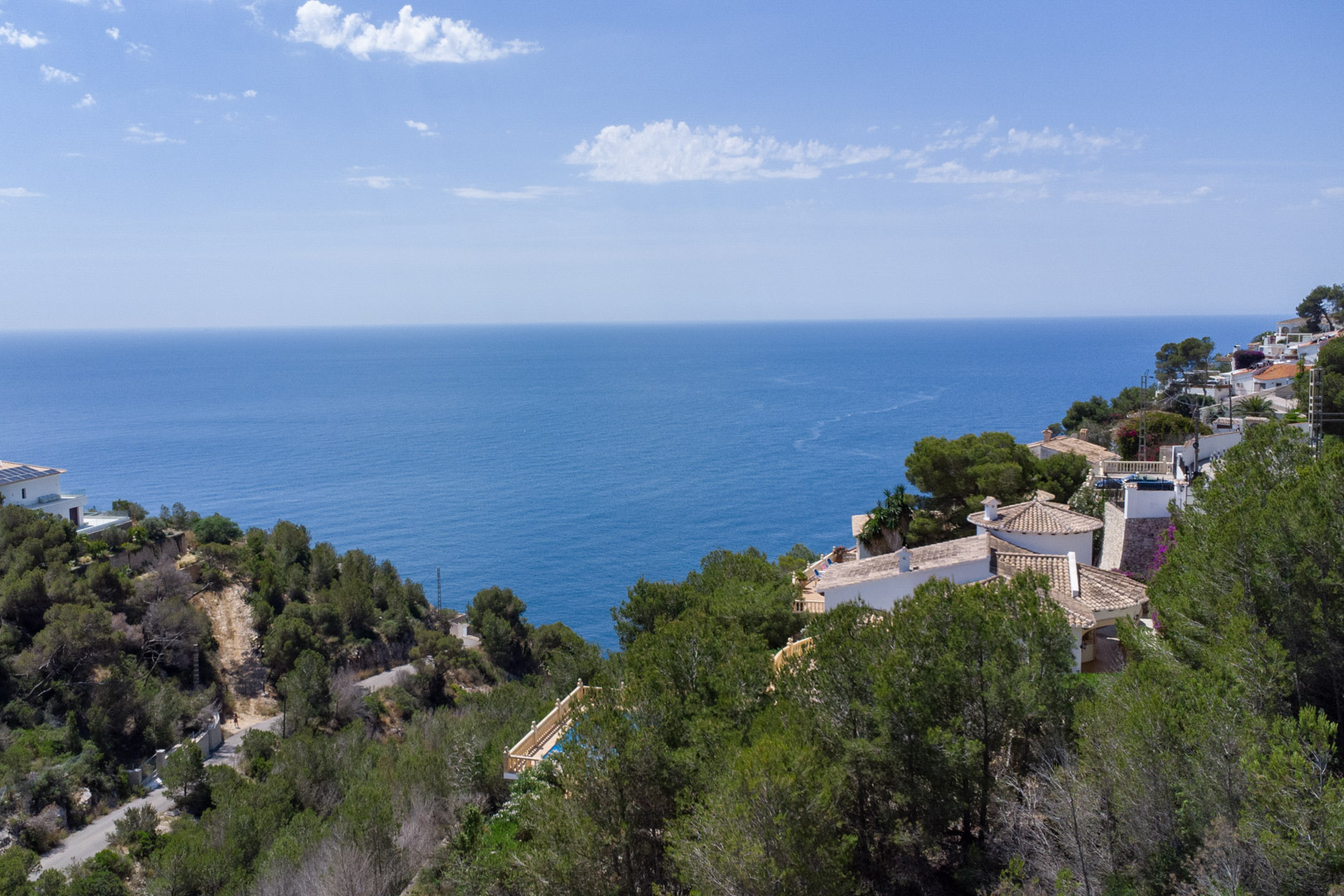Elevated panoramic view of the Mediterranean Sea from the Javea coastline, featuring dense pine forests and traditional villas with terracotta roofs.