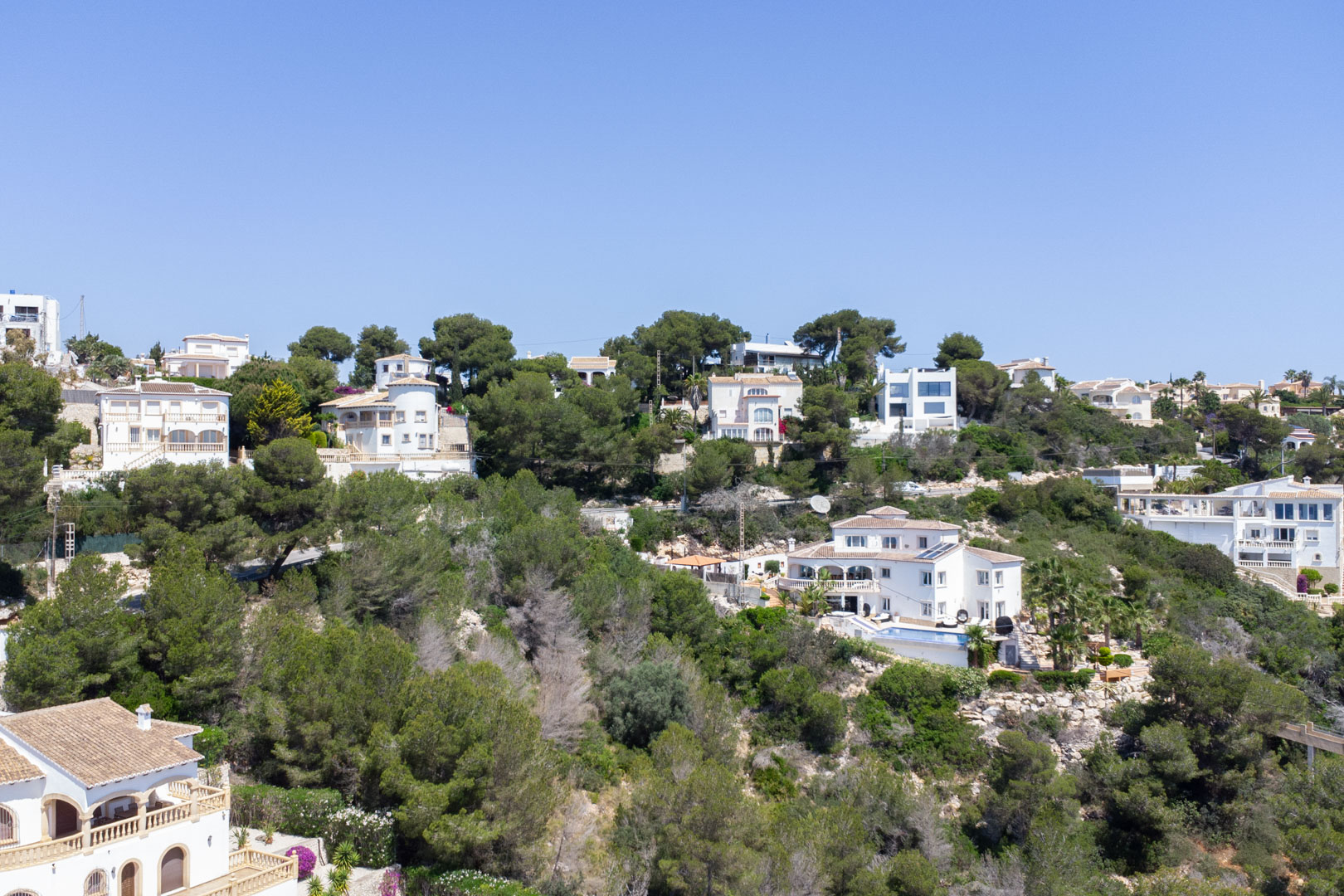 Elevated view of the Costa Nova residential area in Jávea, featuring white villas nestled within a lush Mediterranean pine forest on a sloping terrain.