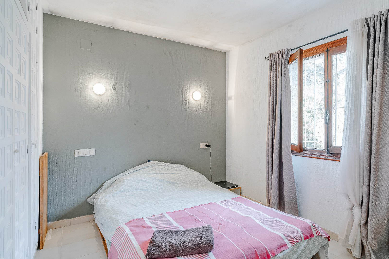 Bedroom featuring a grey accent wall with circular sconces and a built-in white wardrobe. A wooden-framed window provides natural light and neutral curtains.
