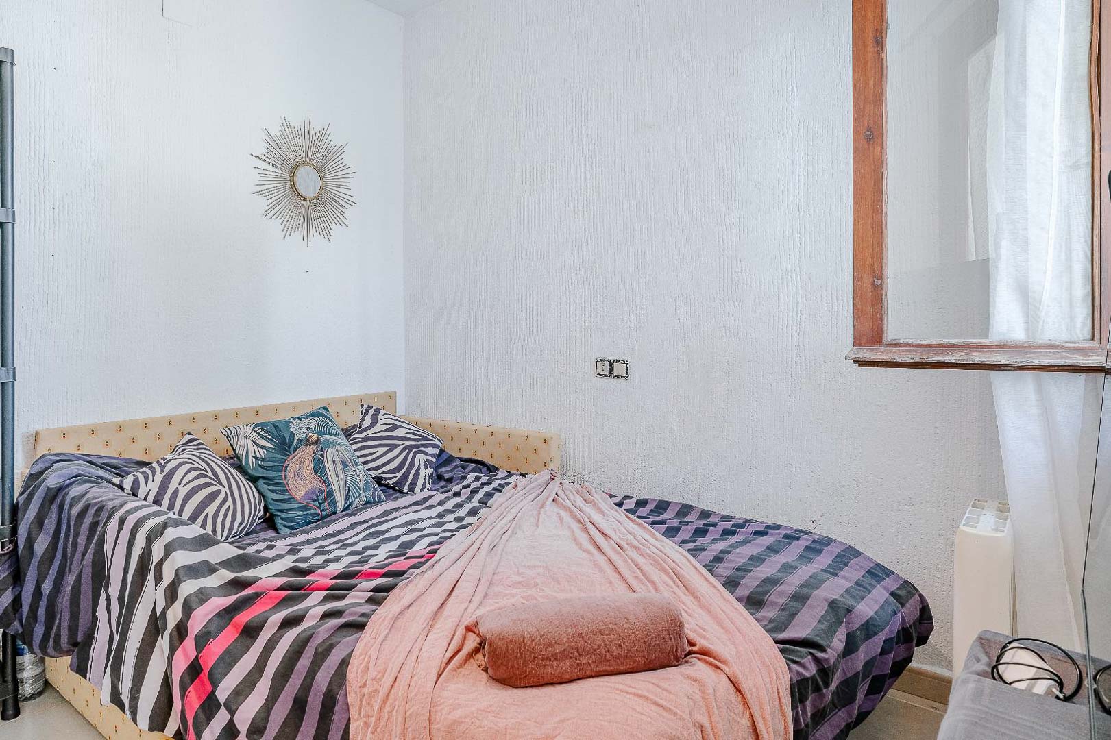 Bright bedroom featuring textured walls, a single bed with a patterned headboard, and a wooden-framed window providing natural light.