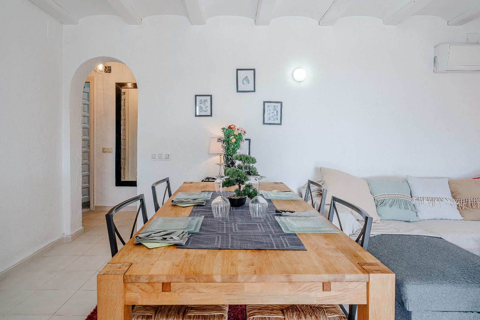 Bright dining area featuring a solid wood table and white beamed ceilings. An arched doorway leads to the hallway, adjacent to the living space with a sofa.