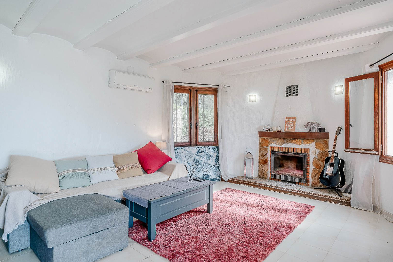 Bright living area featuring white beamed ceilings, a rustic stone fireplace, and split air conditioning. Wooden windows allow ample natural light into the lounge space.