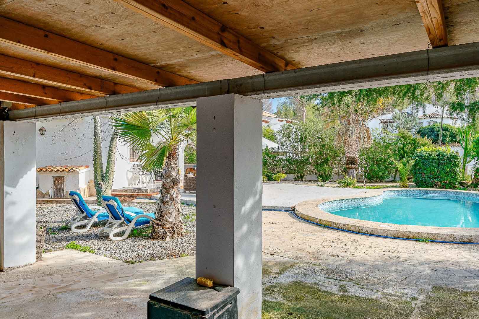 Covered porch featuring exposed wooden beams, overlooking a kidney-shaped swimming pool and a Mediterranean garden with palm trees and loungers.
