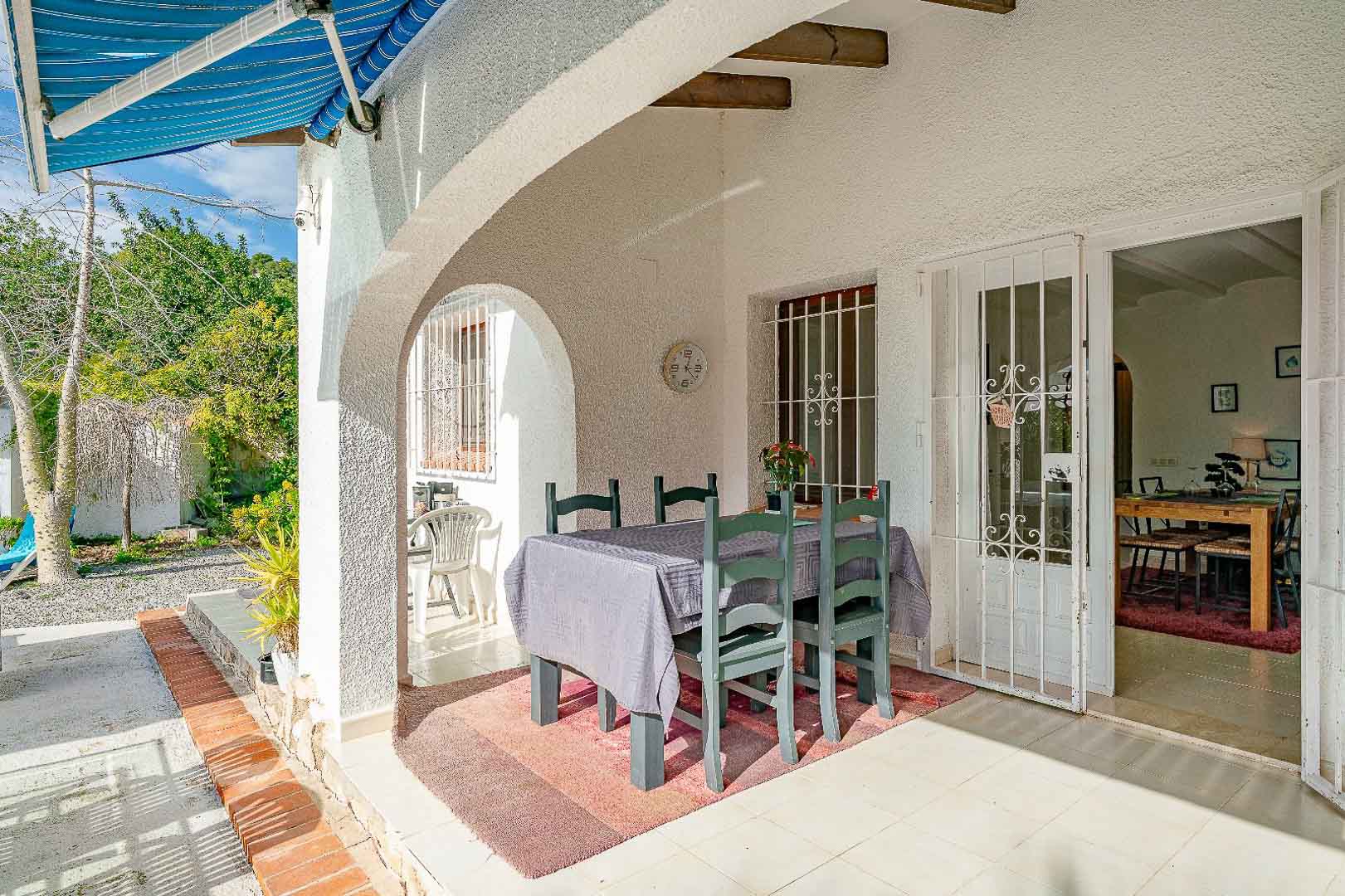 Covered porch featuring Mediterranean arches, wooden beams, and an outdoor dining set. The space connects the garden to the interior via secured glass doors.