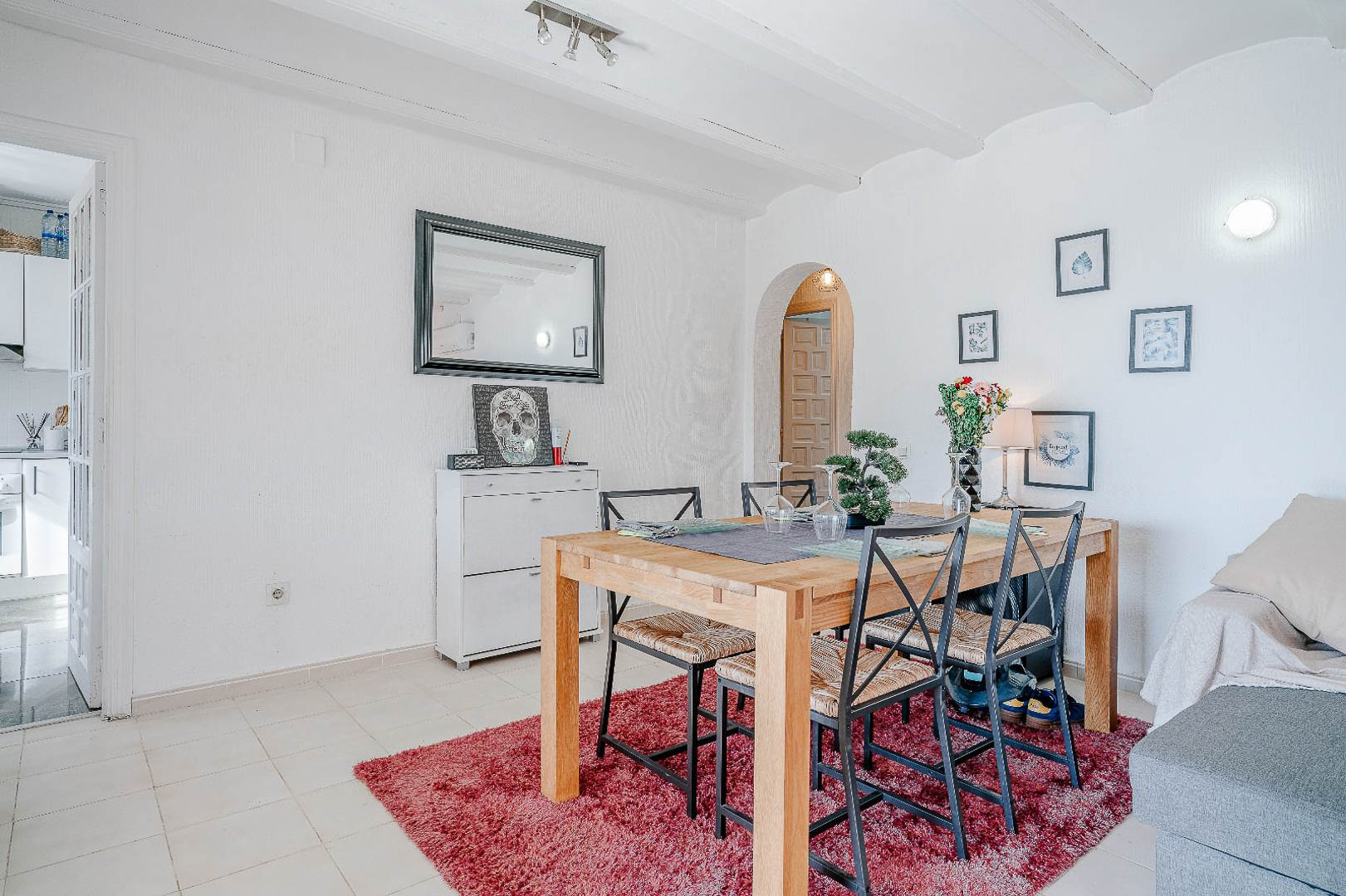 Dining area featuring a solid wood table and metal chairs on a red rug, with white-beamed ceilings and arched doorways connecting to the living space.
