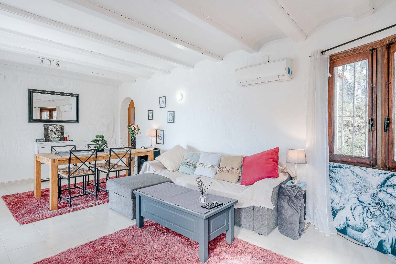 Open-concept living and dining area featuring white exposed ceiling beams, split air conditioning, tiled floors, and a wooden dining set under recessed lighting.