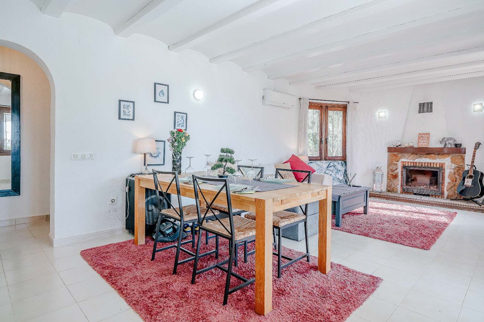 Open-plan living and dining area featuring white beamed ceilings, a natural stone fireplace, and a wooden dining set positioned over red accent rugs.