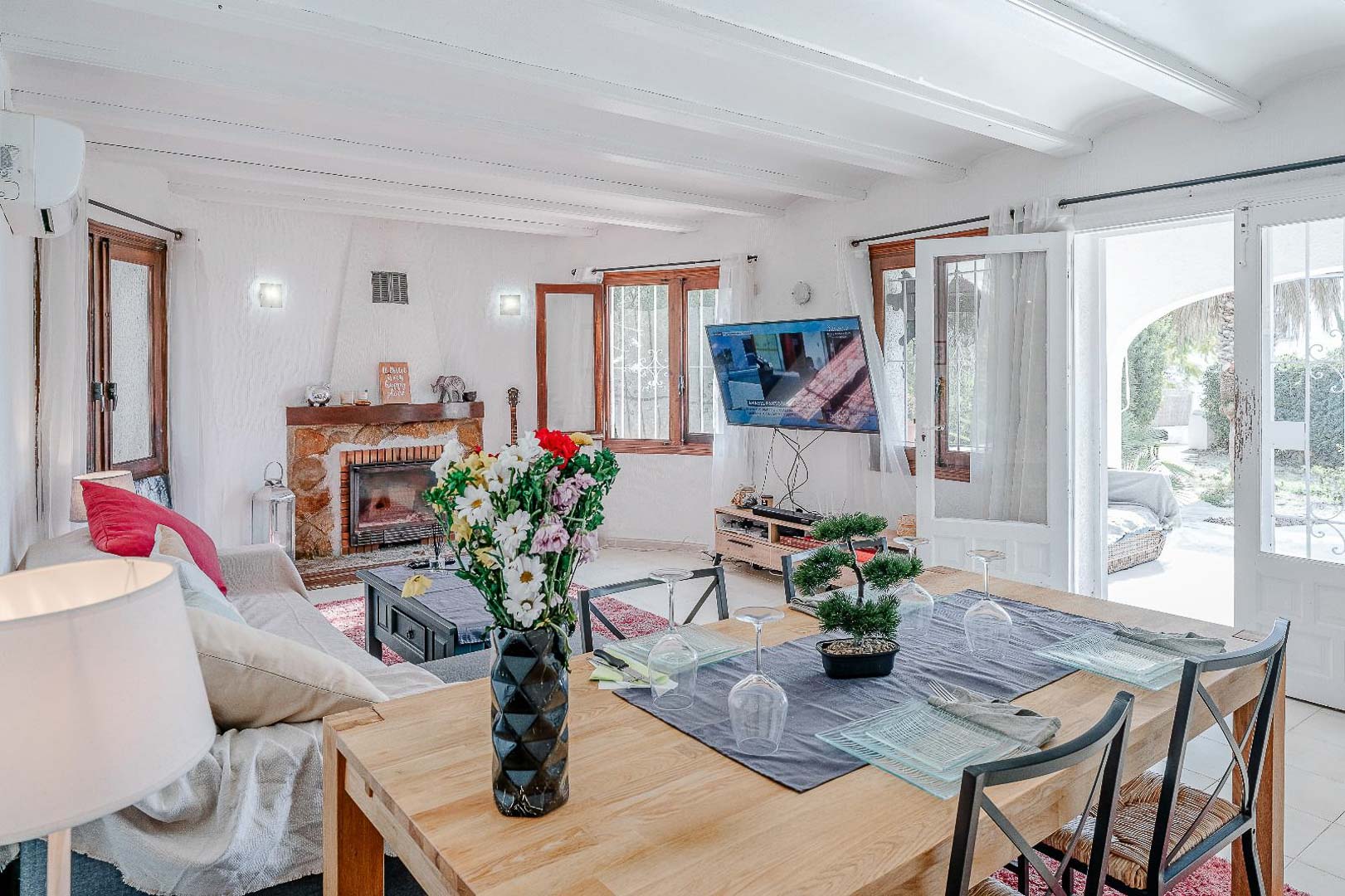 Open-plan living and dining area featuring white beamed ceilings, a traditional stone fireplace, and direct access to the sunlit terrace.