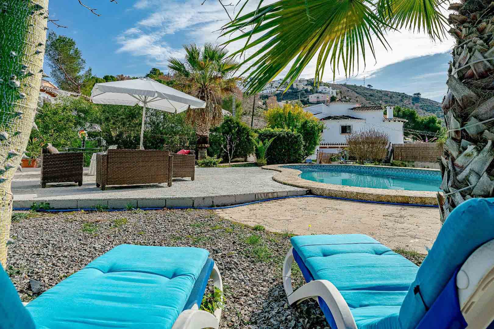 Outdoor pool area featuring a kidney-shaped pool, stone terrace with wicker seating, and blue sun loungers set amidst Mediterranean palms and gravel landscaping.