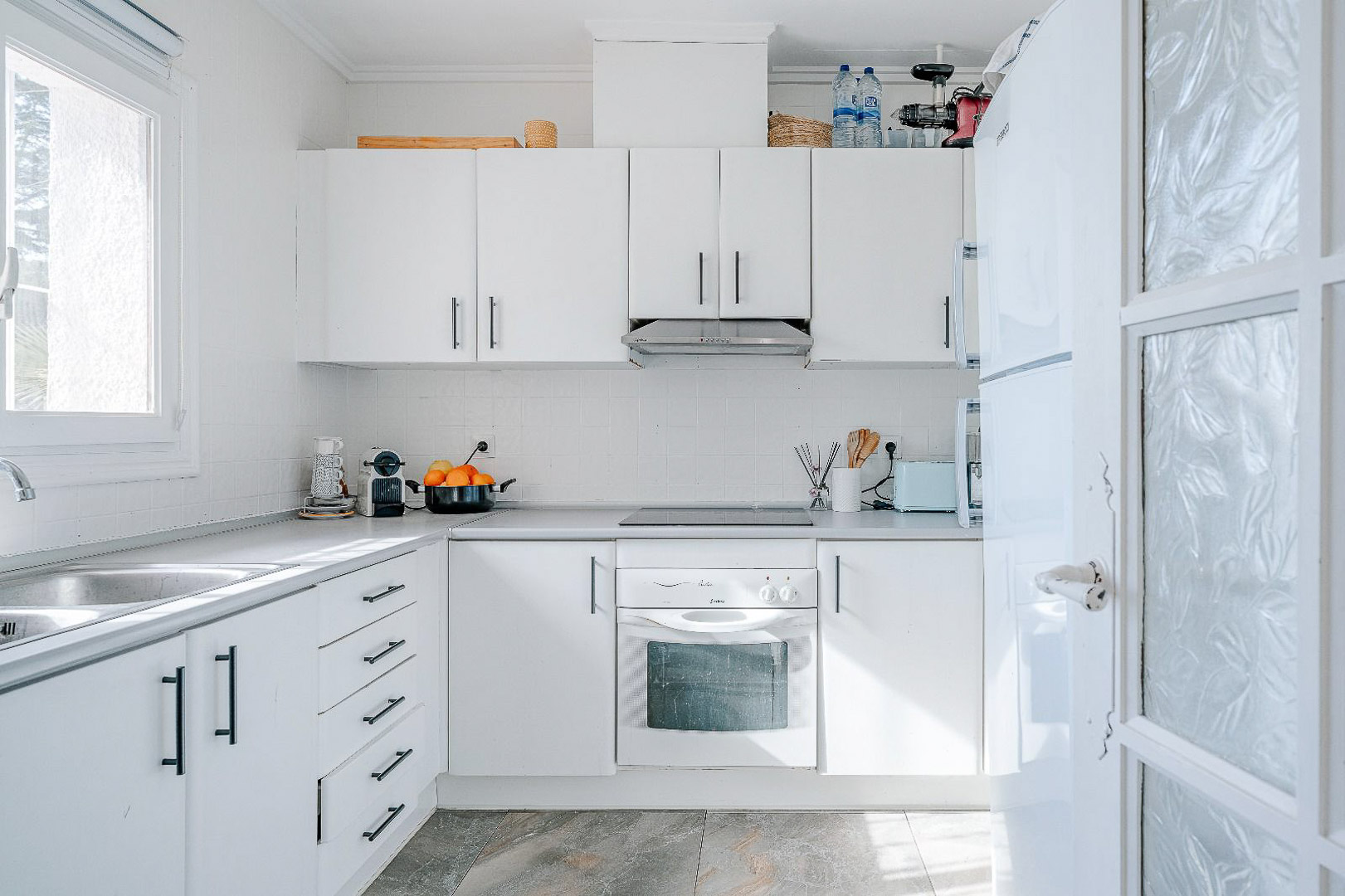 Practical white kitchen featuring black handles, integrated appliances, and a double stainless steel sink positioned under a window for optimal natural lighting.