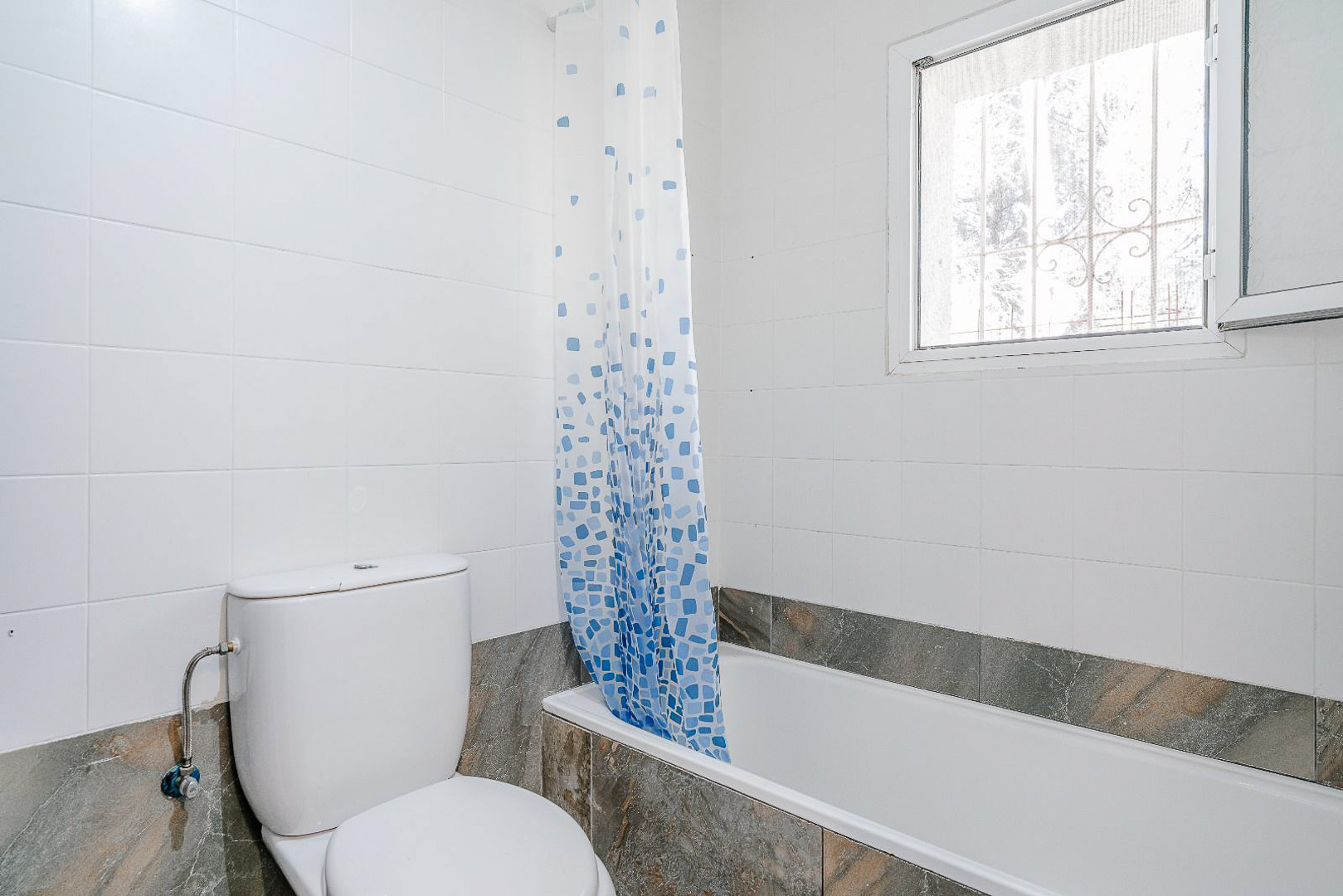 Renovated bathroom featuring a built-in bathtub with marble-effect tiling and a white ceramic toilet. A window with a security grate provides natural light.