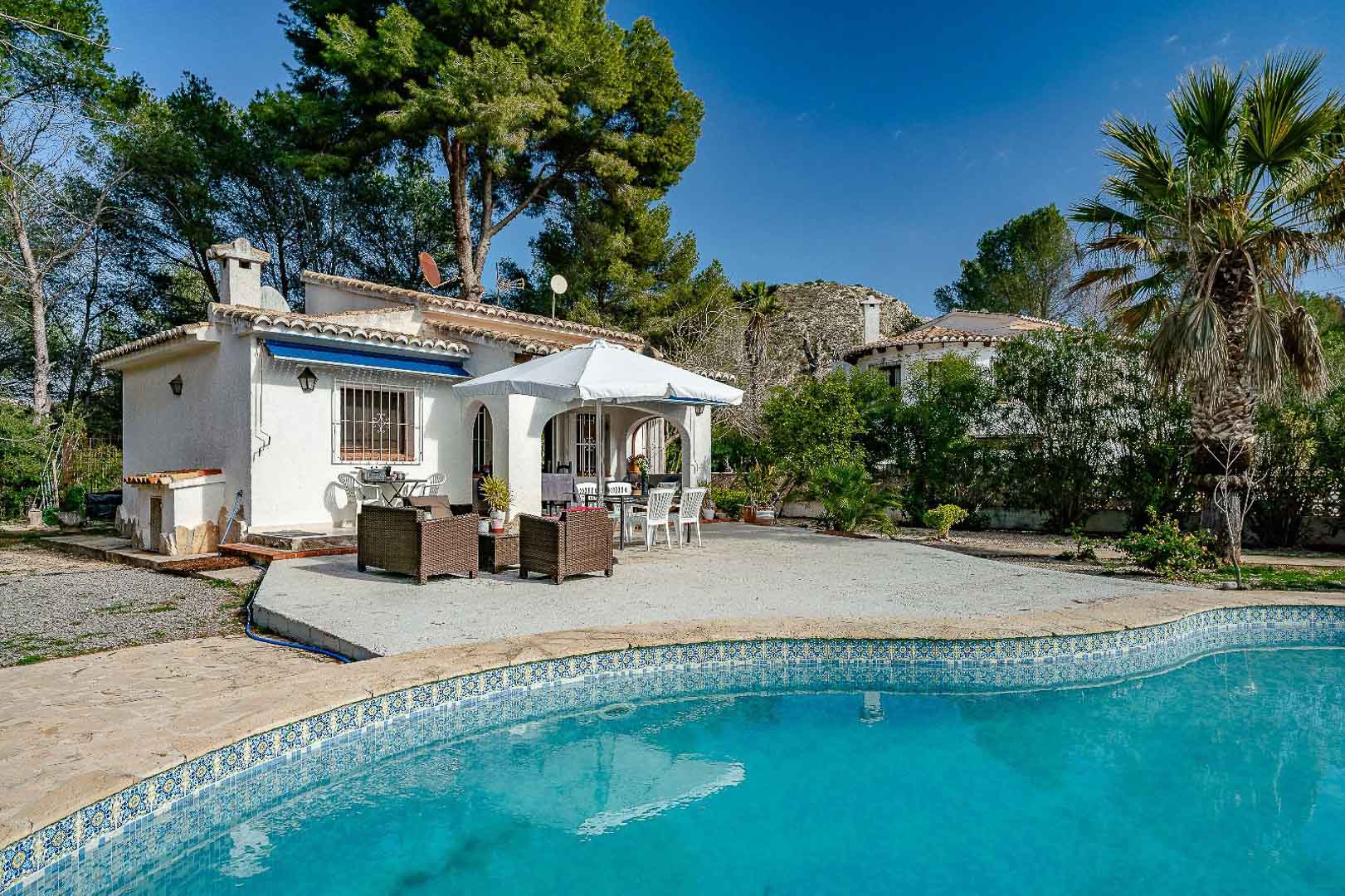 Villa exterior featuring a blue mosaic tile pool, spacious paved terrace with outdoor seating, and a traditional arched porch under a clear blue sky.