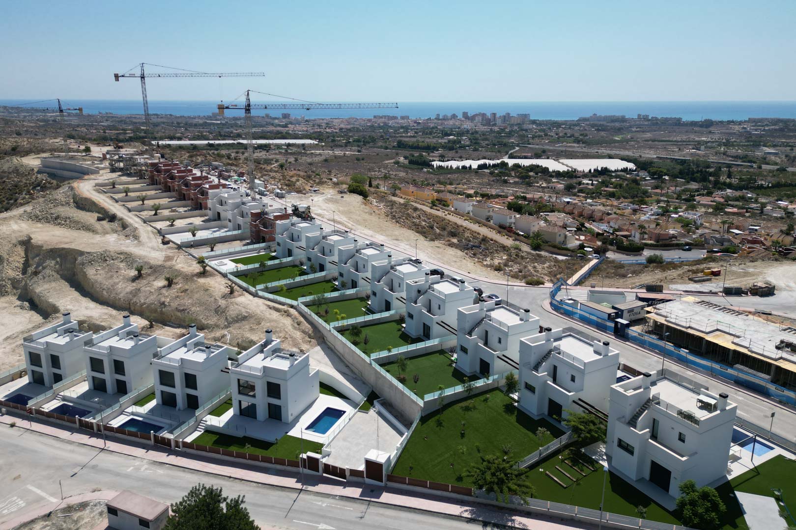 Aerial perspective of modern white villas on a hillside, showing private garden plots, individual swimming pools, and an elevated view of the Mediterranean coastline.