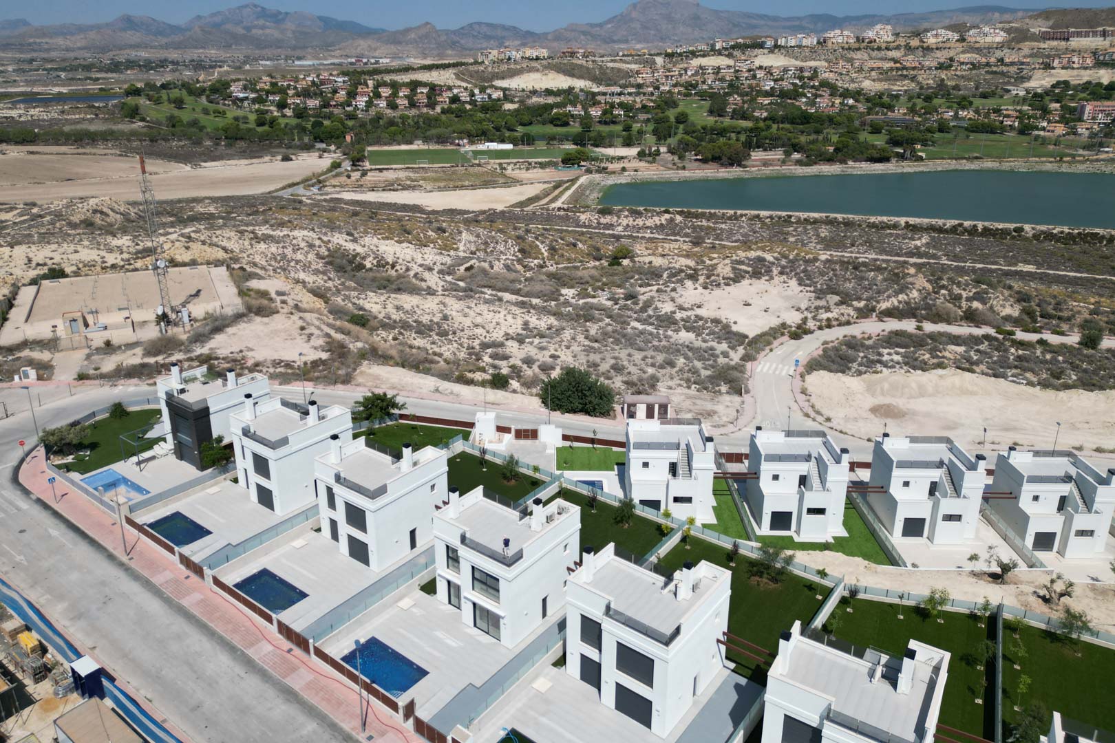 Aerial view of contemporary white villas featuring private pools and gardens, located near a golf course and reservoir with mountain views.