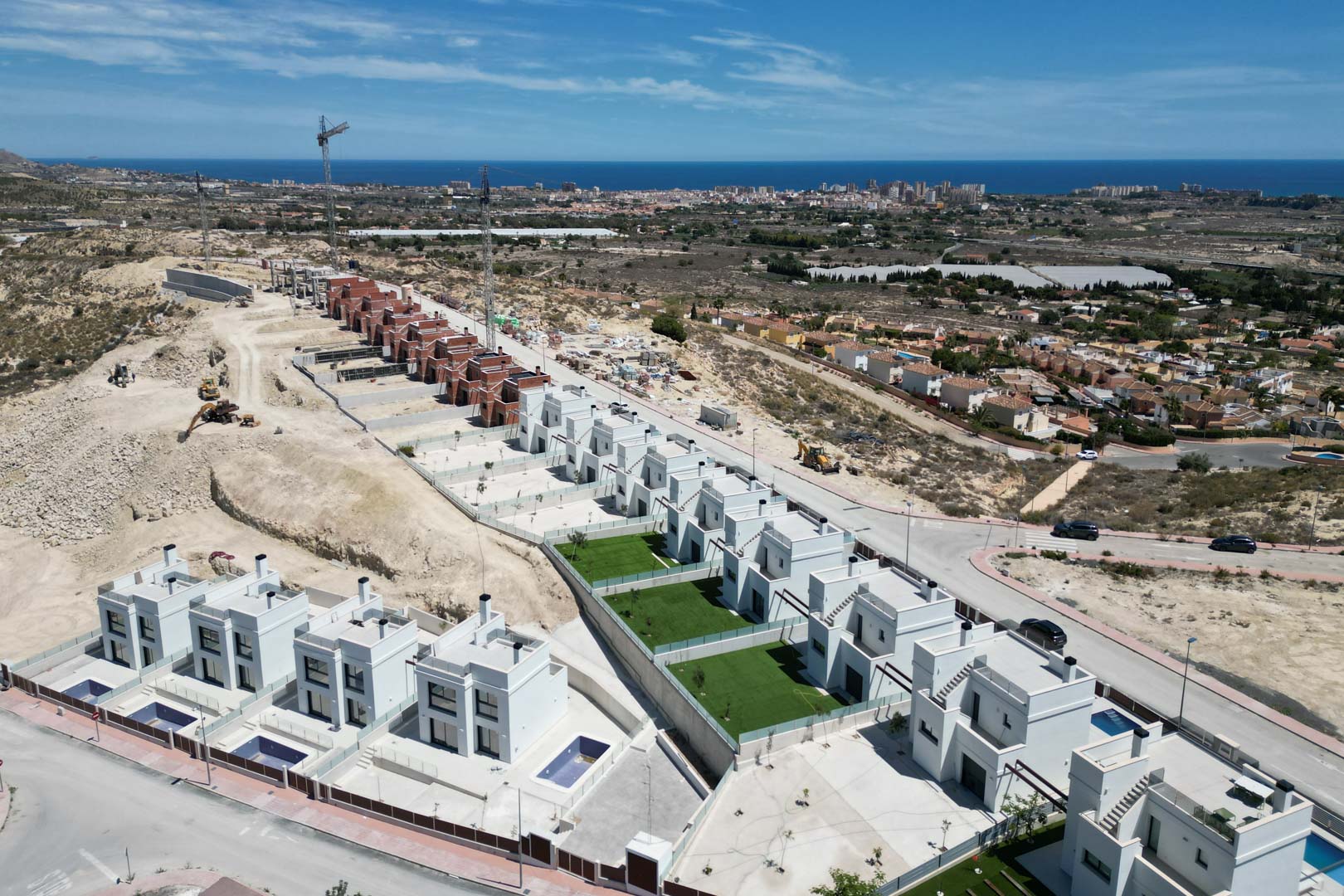 Aerial view of contemporary white villas on a hillside, featuring private pools and gardens with panoramic views towards the Mediterranean Sea and coastal skyline.