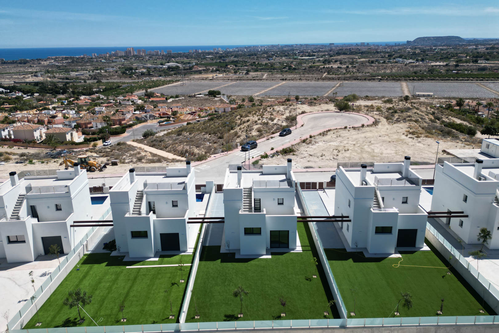Aerial view of modern white villas featuring private lawns, flat roof terraces, and minimalist architecture overlooking the Mediterranean coastline and city skyline.