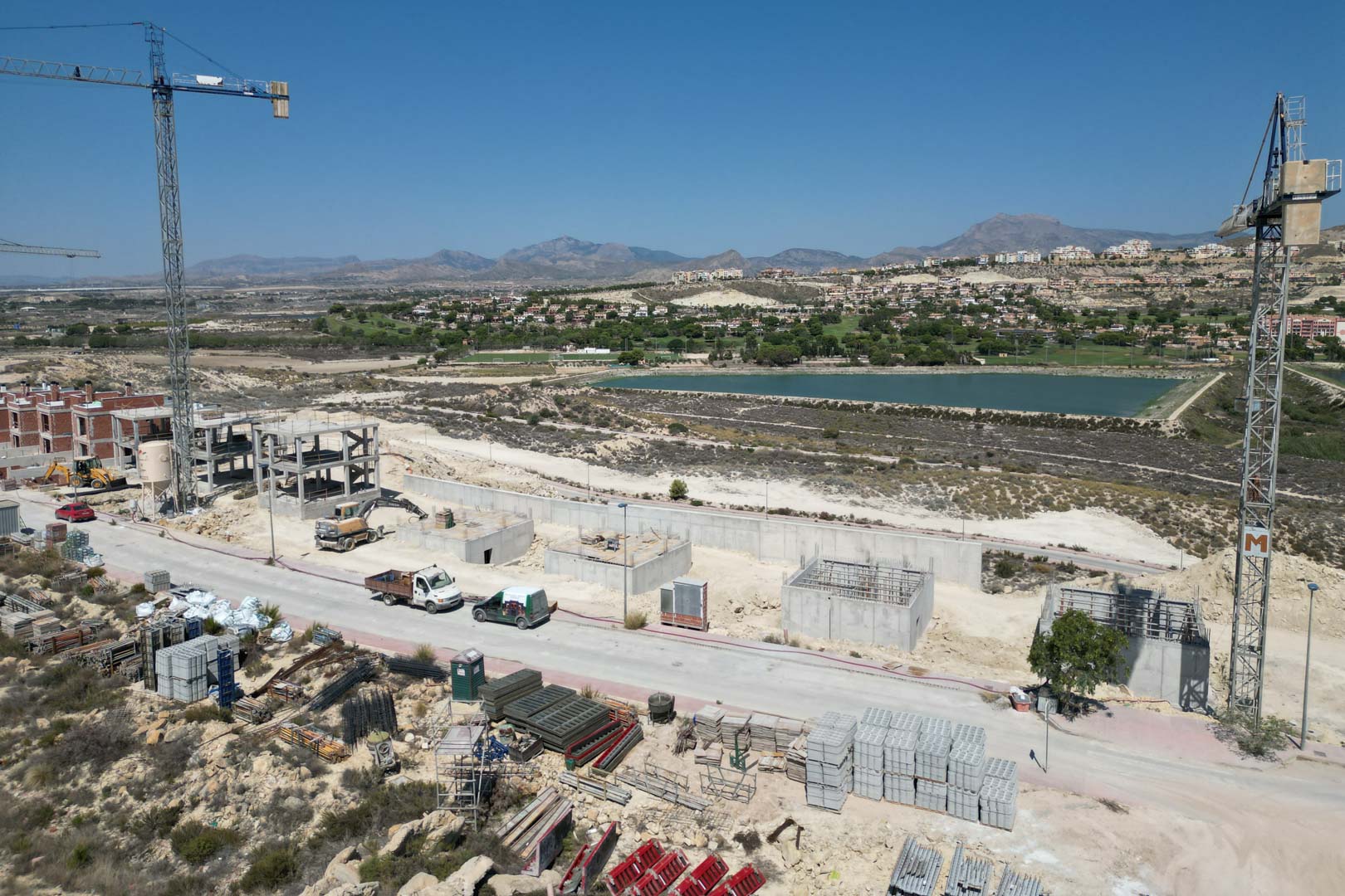 Aerial view of the villas under construction, featuring concrete structures, cranes, and a scenic backdrop of the nearby lake and mountain range.
