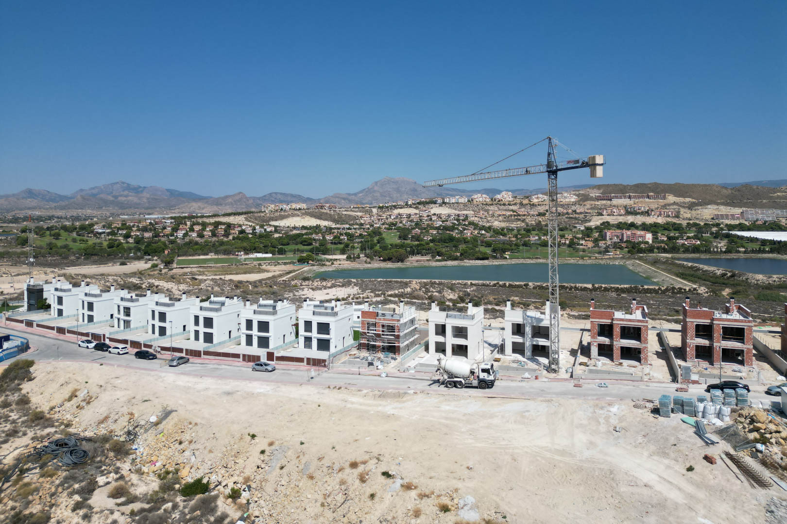 Aerial view showing the construction progress of contemporary villas, featuring finished white facades and brick structures alongside a crane and mountain backdrop.