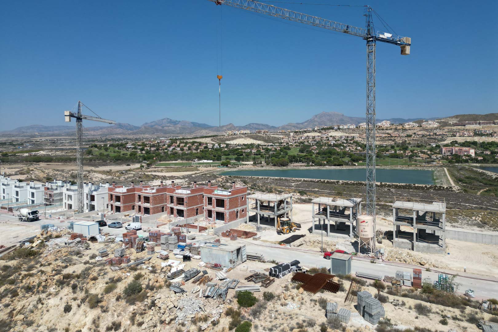 Aerial view showing the construction progress of the villas, featuring brick and concrete shells, cranes, and machinery set against a golf course and mountain backdrop.