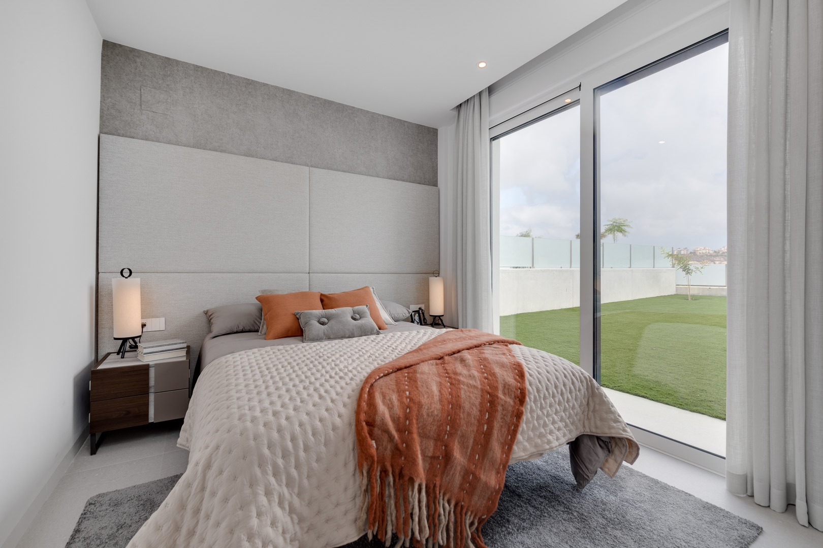 Contemporary bedroom featuring a padded headboard, earth-toned textiles, and floor-to-ceiling glass doors opening directly onto a private lawn.