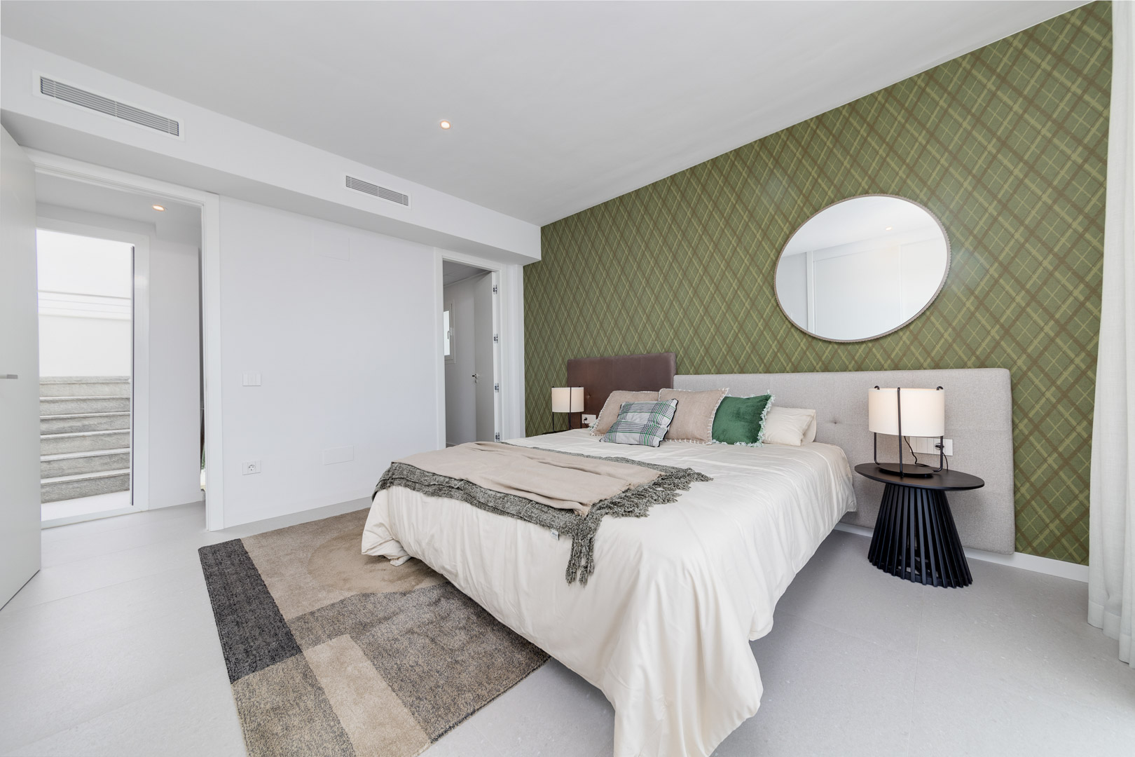 Contemporary bedroom featuring an olive green patterned accent wall, circular mirror, and integrated climate control vents above neutral-toned flooring.