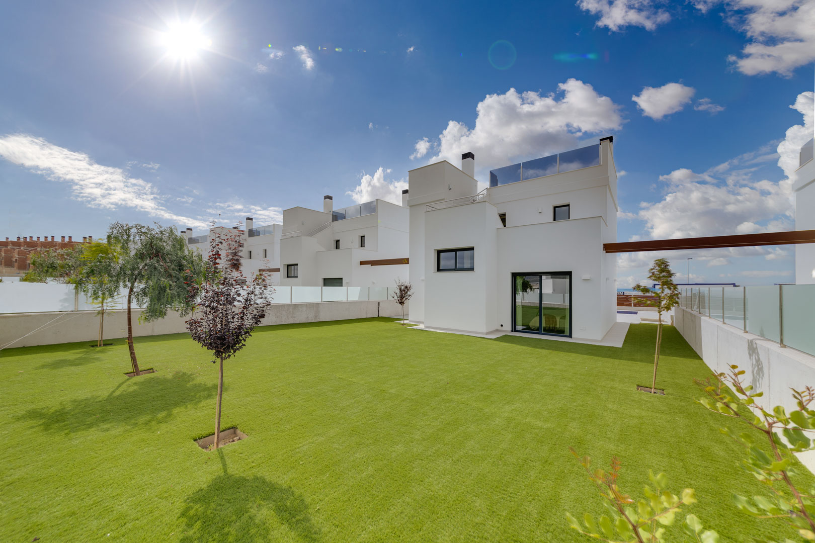 Exterior view of the villa featuring a minimalist white facade, a spacious lawn with young trees, and rooftop glass balustrades.