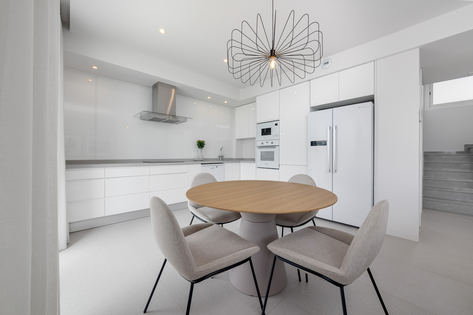 Minimalist white kitchen featuring integrated appliances, grey countertops, and a dining area with a round wooden table and wire pendant light.