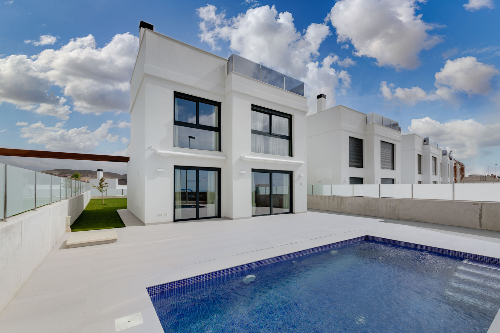 Modern white villa exterior featuring black window frames, a spacious tiled terrace, and a private swimming pool with blue mosaic tiling.