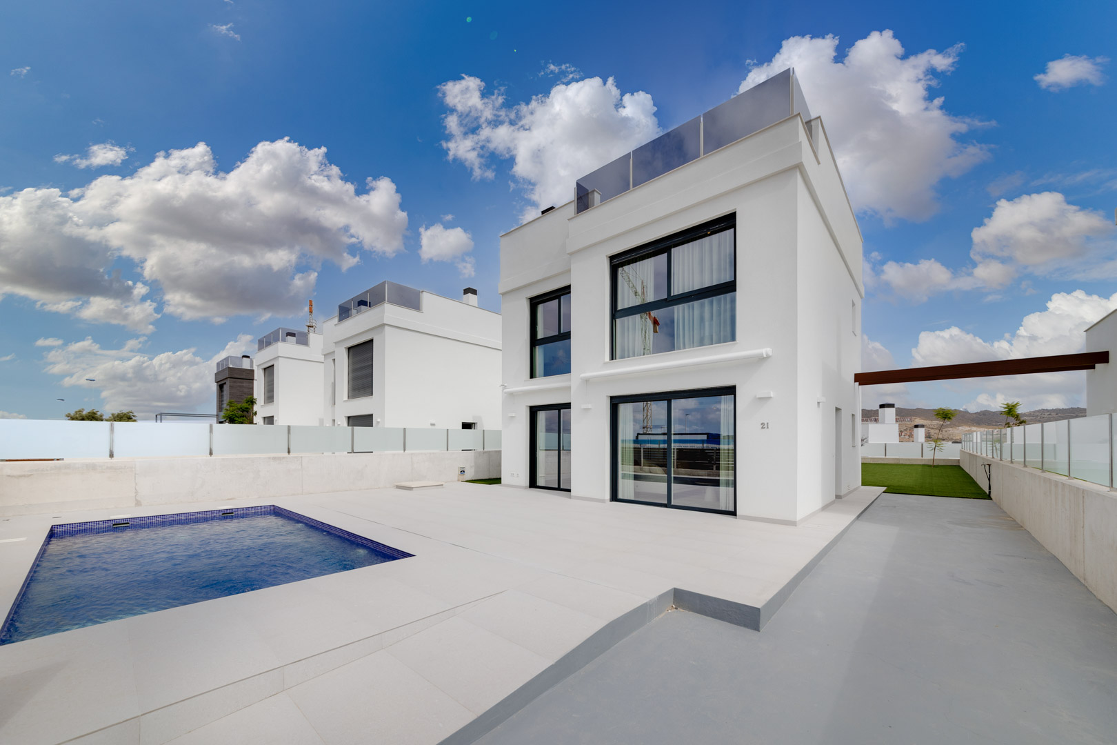 Modern white villa featuring a private rectangular pool, expansive tiled terrace, and large minimalist windows under a bright sky.