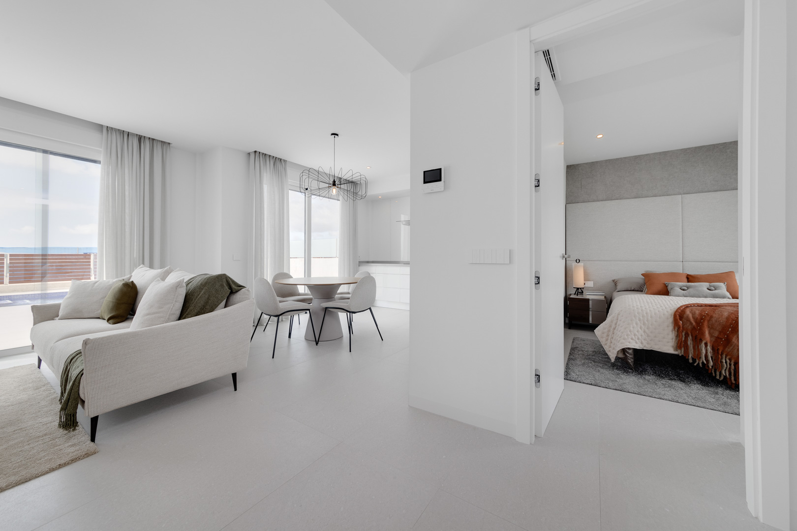 Open-concept living and dining area featuring large-format tiling, designer lighting, and a view into a minimalist bedroom with terracotta accents.