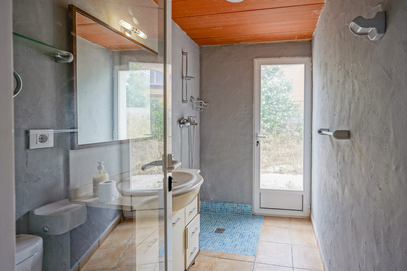 Bathroom featuring textured grey walls, an orange slatted ceiling, and a blue mosaic tile shower floor. A glass door provides direct access to the garden area.