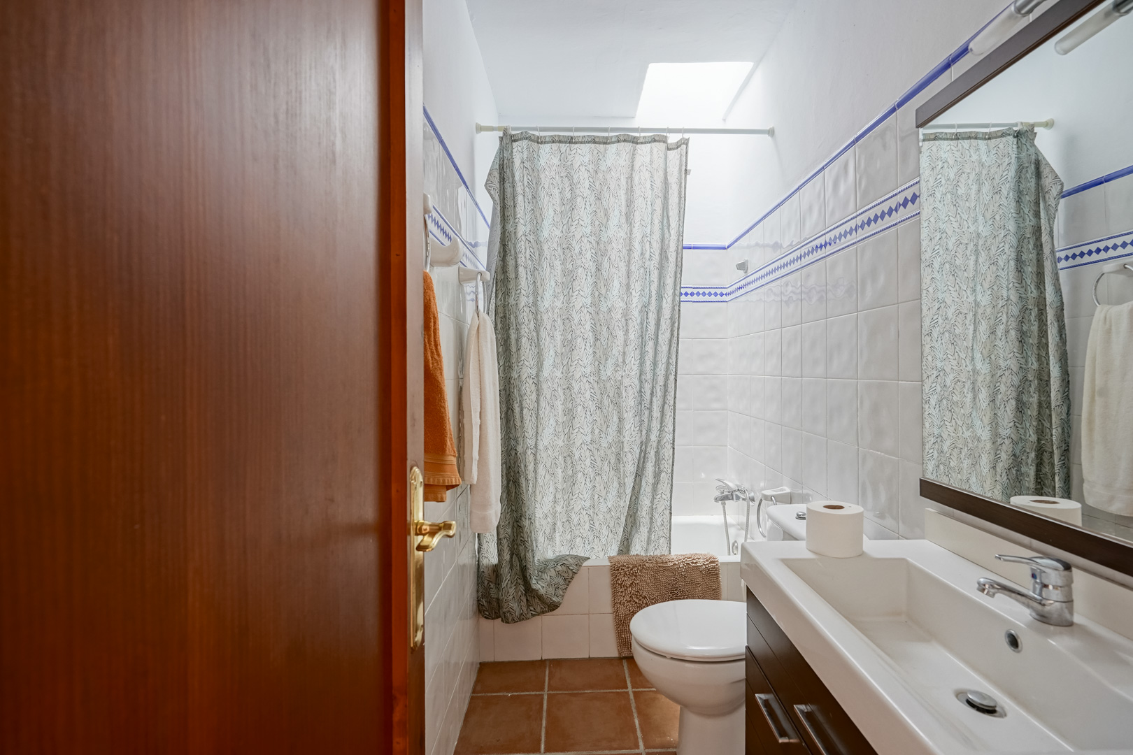 Bathroom featuring white wall tiles with blue accents, a modern vanity with dark cabinetry, and a bathtub illuminated by a natural skylight.