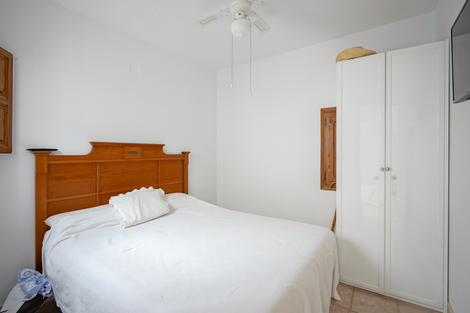Bedroom featuring a double bed with a wooden headboard, a white wardrobe, and a ceiling fan. White walls and tiled floors enhance the natural light.