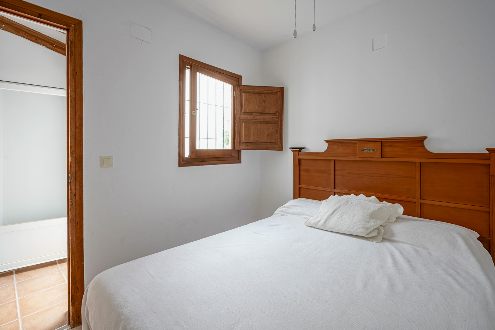Bedroom featuring a solid wood headboard, a traditional shuttered window with security bars, and an adjacent room with terracotta tiling.