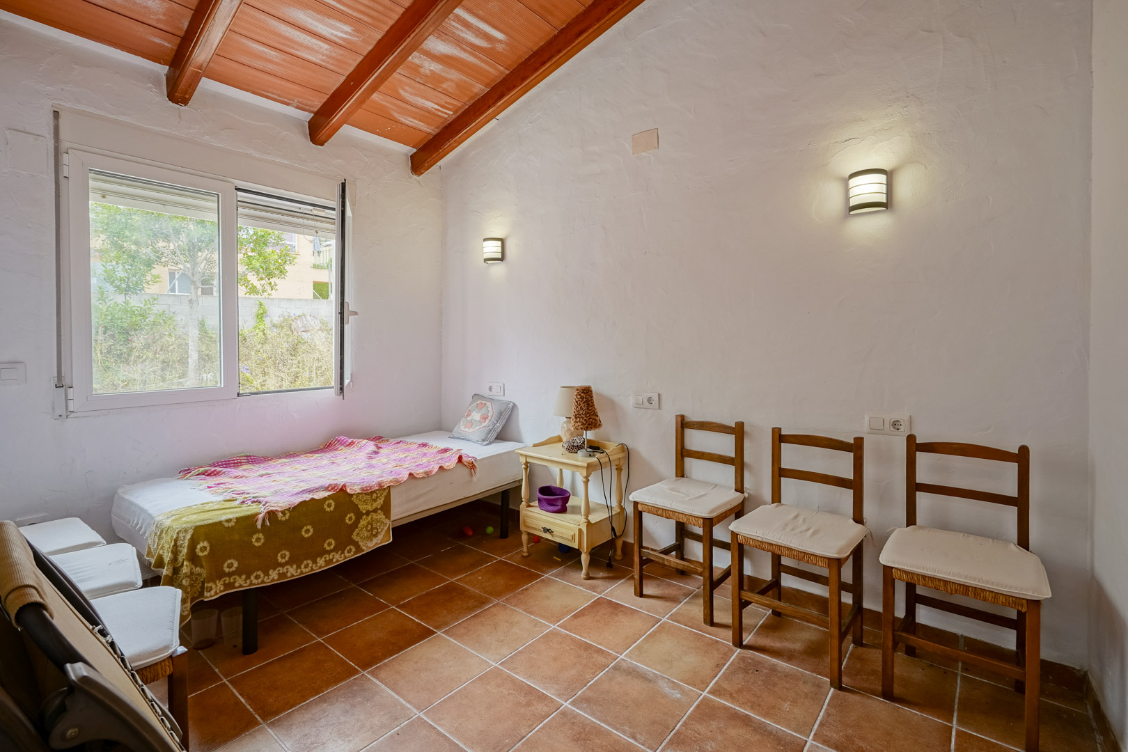 Bedroom featuring slanted ceilings with exposed wooden beams, terracotta tile flooring, and white textured walls. Includes wall-mounted lighting and a window.