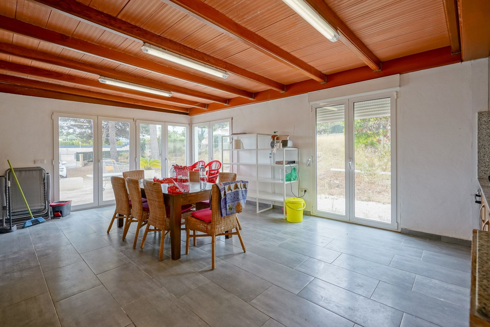 Bright dining area featuring an exposed wood-beam ceiling, grey tiled flooring, and large white-framed glass doors providing direct access to the garden.