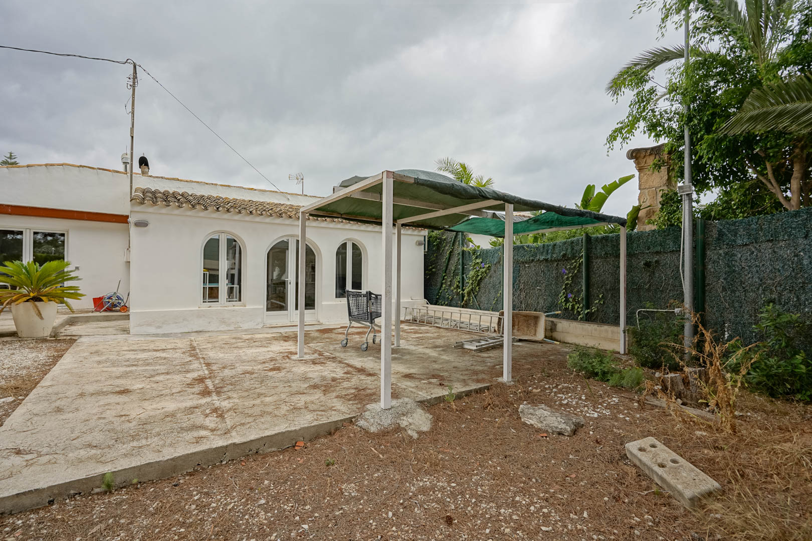 Exterior view of the existing house featuring white walls, arched windows, and a concrete patio area with a temporary pergola structure and perimeter greenery.