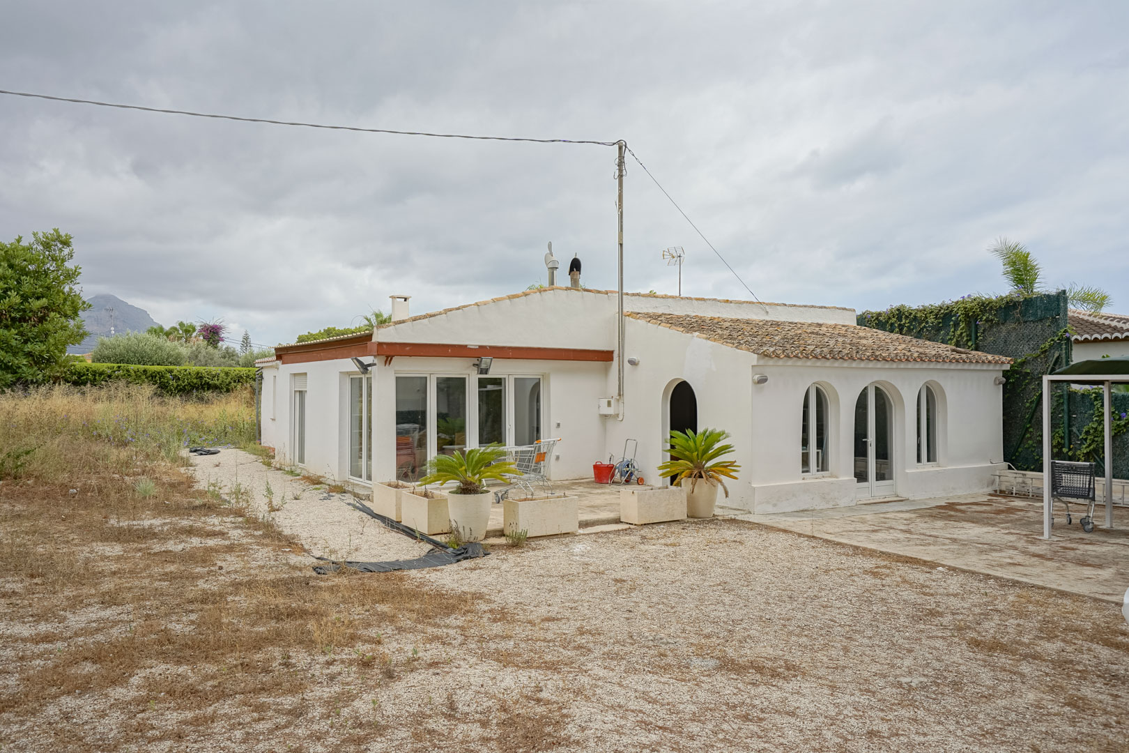 Exterior view of the Mediterranean villa featuring traditional arches, white joinery, and a tiled roof, set on a spacious flat plot with mountain silhouettes.