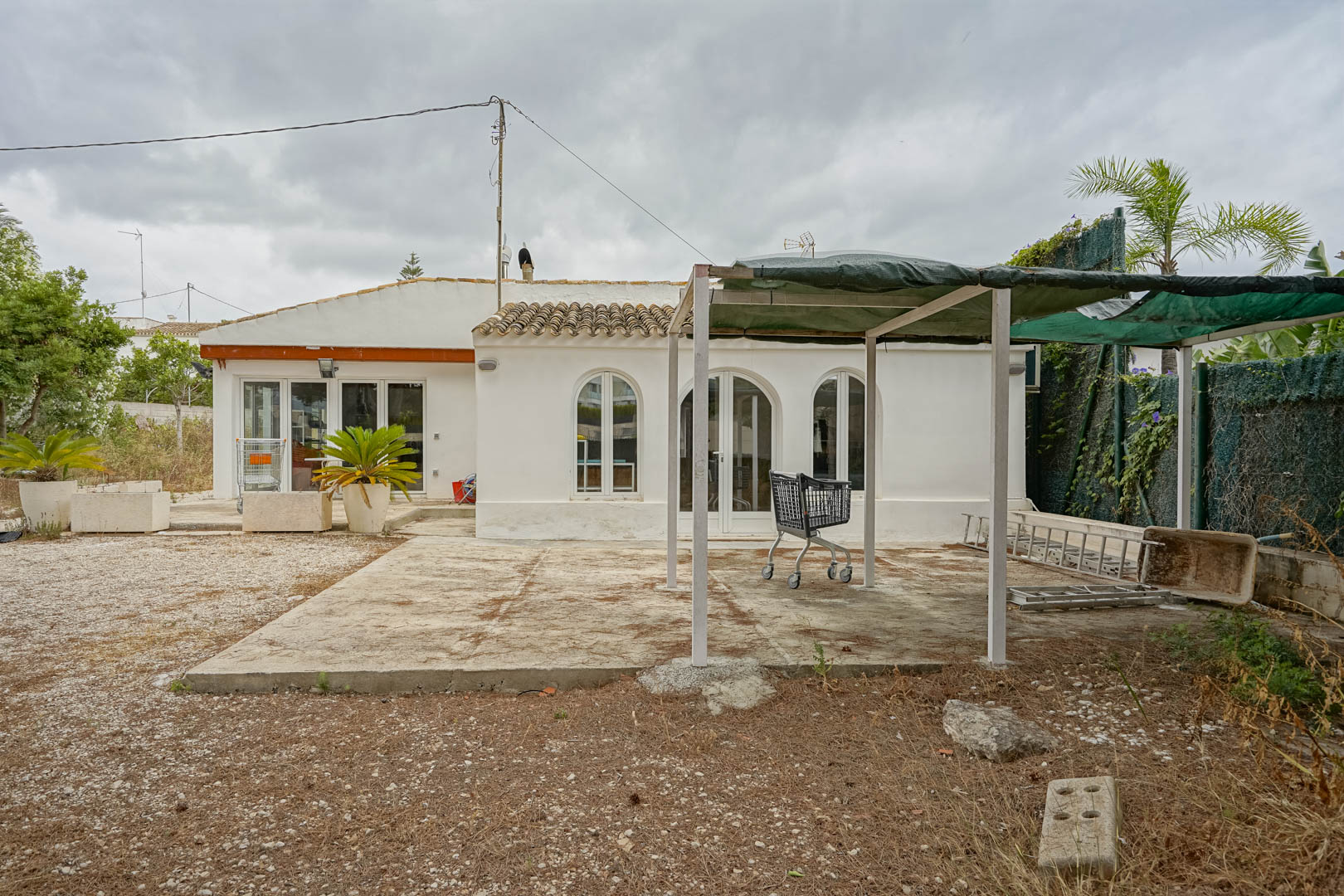 Exterior view of the single-story villa featuring arched windows, a gravel yard, and a metal pergola structure with shade cloth.