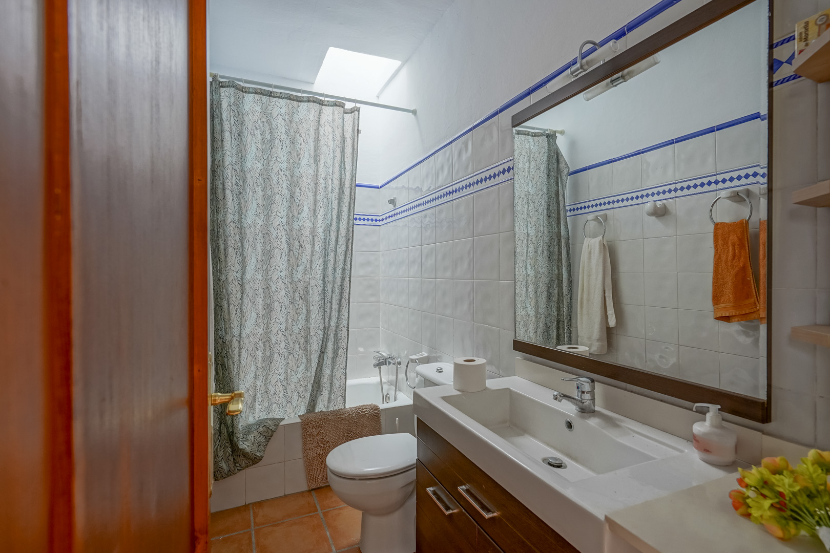 Functional bathroom featuring white tiling with blue accents, a dark wood vanity, and a bathtub positioned under a natural skylight.