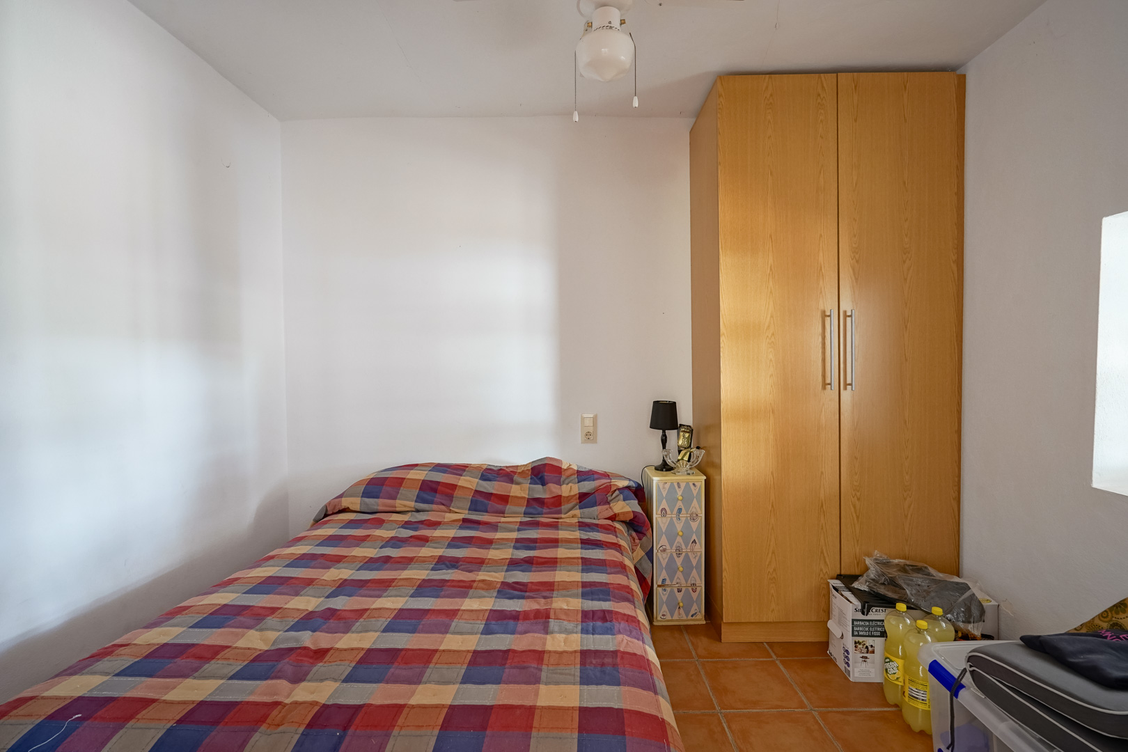 Functional bedroom featuring terracotta tile flooring, a tall wooden wardrobe, and white walls that enhance the natural light.