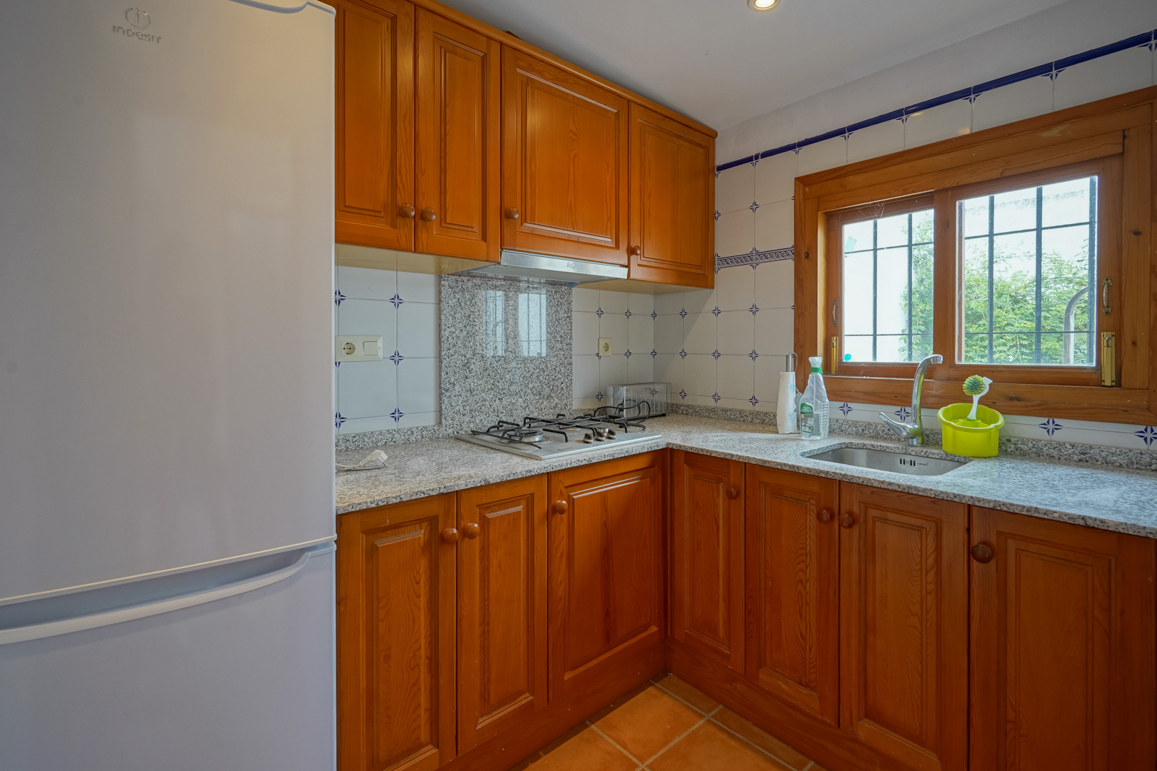 Functional kitchen featuring solid wood cabinetry, grey granite countertops, and a gas hob. A wooden-framed window provides natural light over the stainless steel sink.