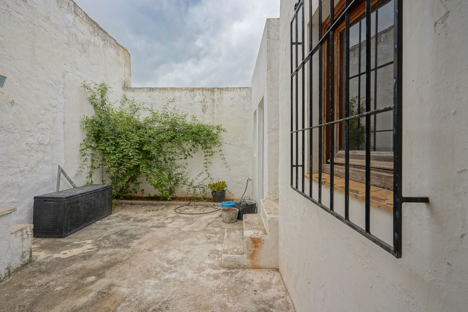 Interior patio featuring whitewashed walls, concrete flooring, a window with traditional iron security bars, and an outdoor storage area.