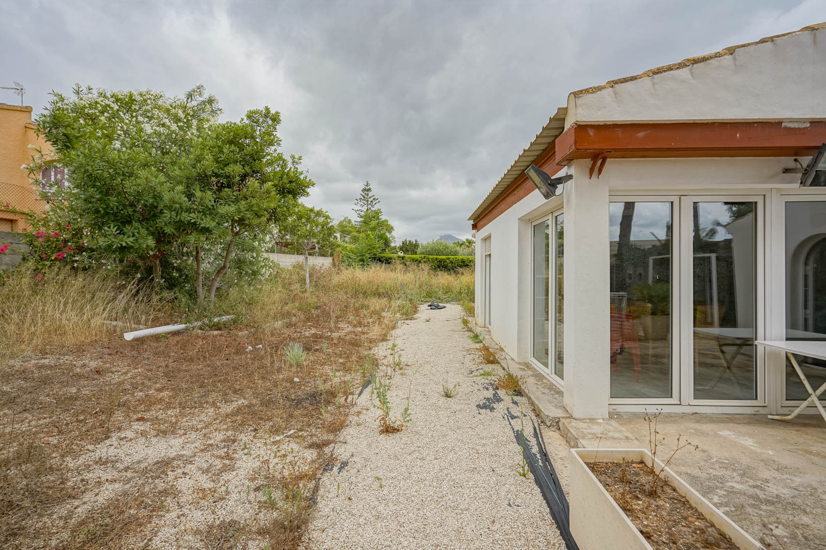 Side facade of the villa featuring large white PVC windows and direct access to an expansive natural plot with Mediterranean vegetation.