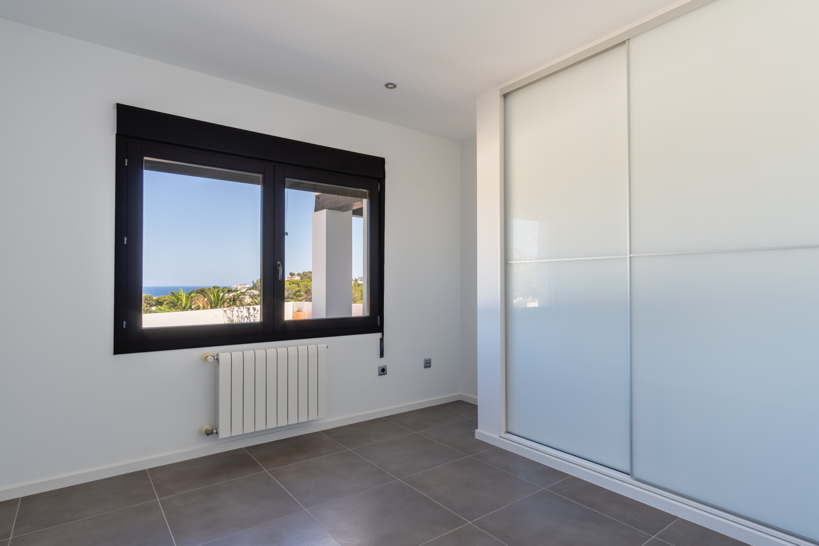Bright bedroom featuring built-in wardrobes with sliding doors, a radiator, and a dark-framed window offering views of the Mediterranean Sea and coastline.
