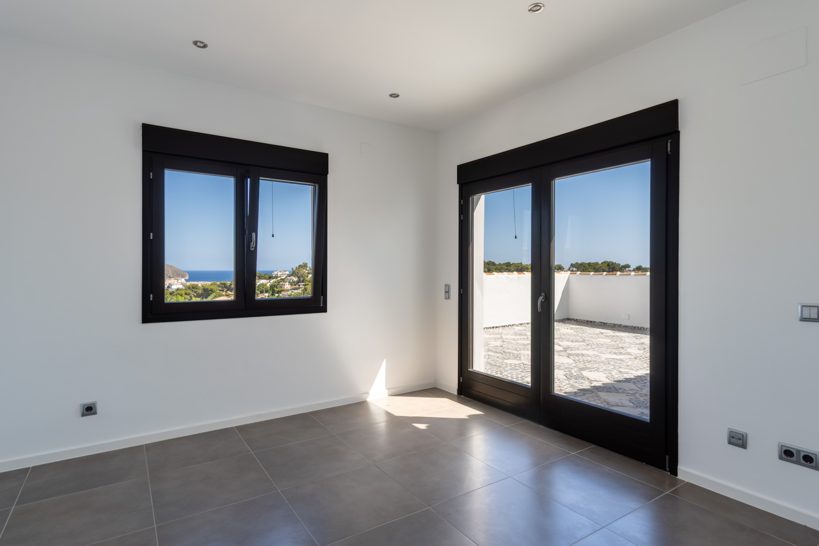 Bright room featuring grey ceramic tiles, dark-framed glazing, and direct access to a tiled terrace with panoramic views of the Mediterranean Sea.