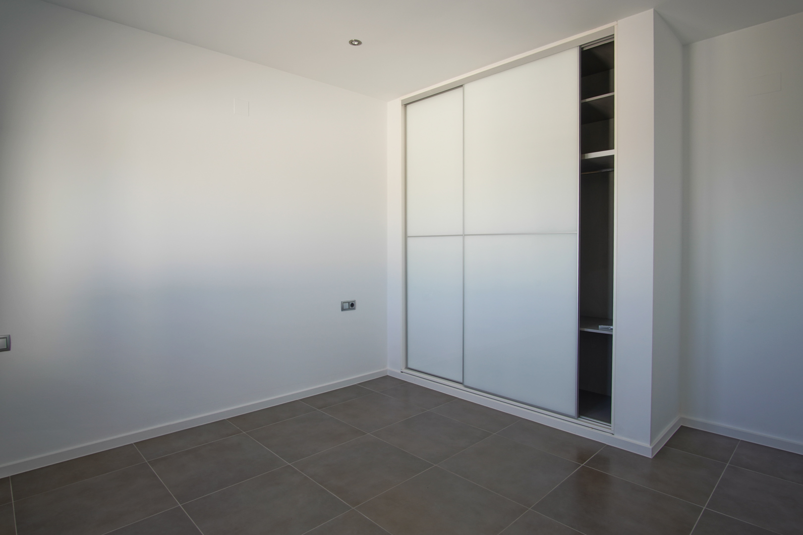 Built-in wardrobe featuring white sliding doors and internal shelving, set against minimalist white walls and large-format grey floor tiles.