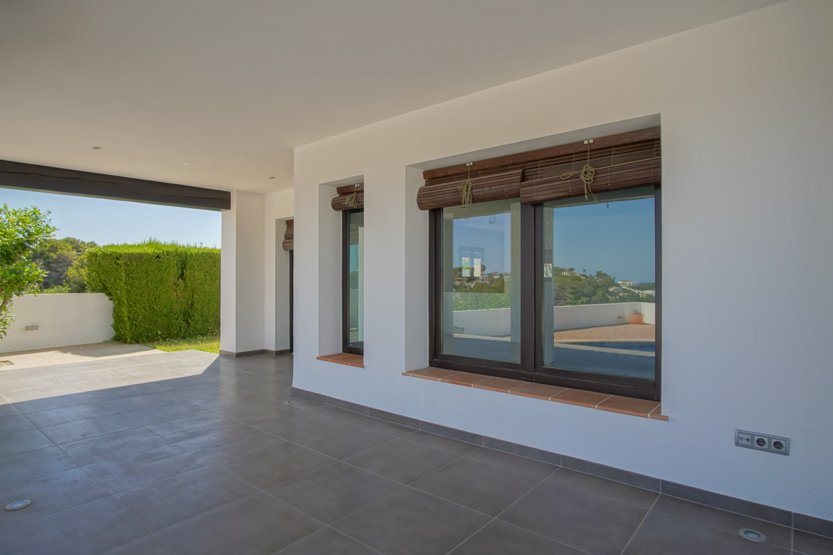 Covered porch featuring grey tiled flooring, white walls, and wood-effect aluminum windows with bamboo roll-up blinds and terracotta sills.