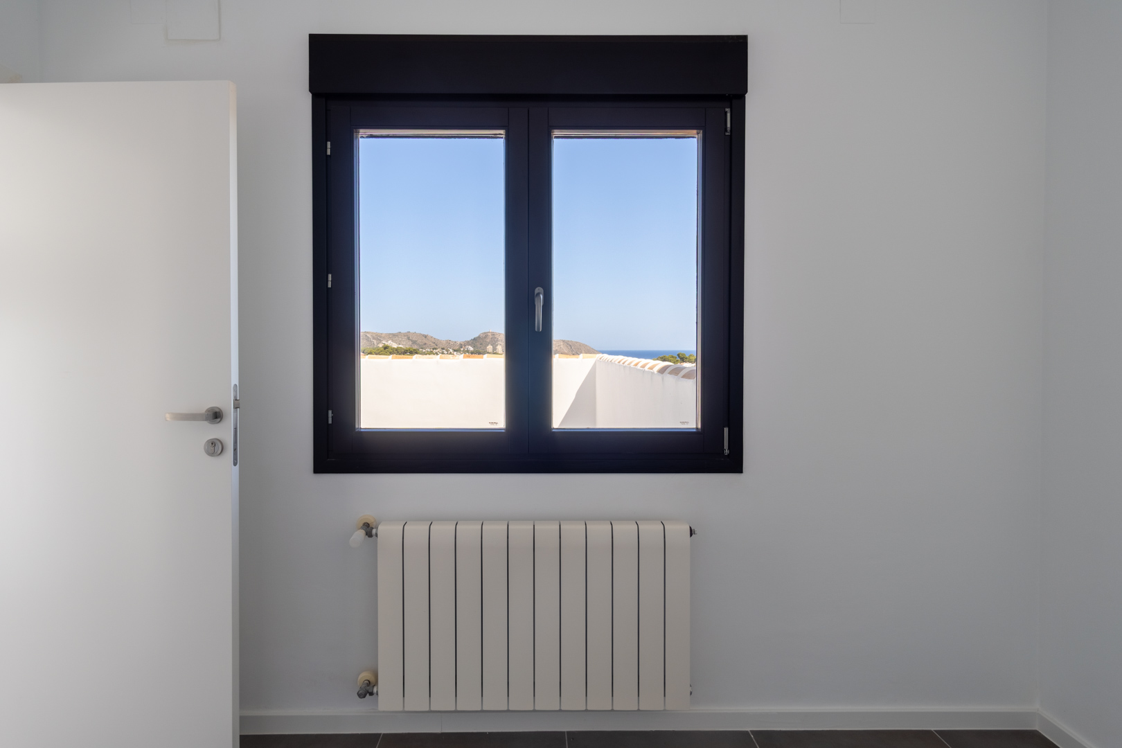 Dark-framed aluminum window with double glazing framing views of the sea and Cabo de Moraira, positioned above a white panel radiator in a bright, minimalist interior.