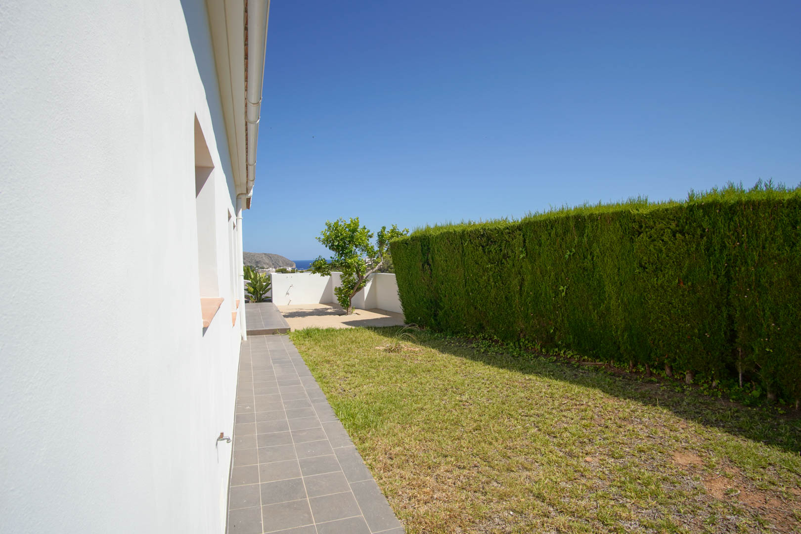 Exterior side walkway with grey tiling, a lawn area, and a tall privacy hedge, offering a glimpse of the sea in the distance under a clear blue sky.